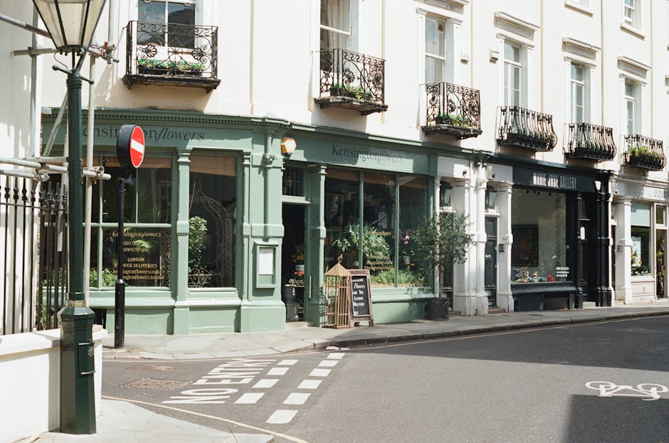 A storefront of a florist shop with large display windows showcasing colorful floral arrangements inside. The building has a soft green facade with decorative wooden framing, and a small chalkboard si