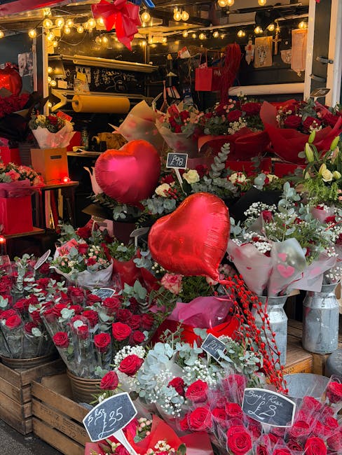 A floral display featuring numerous red and white roses, carnations, and baby's breath arranged in baskets and bouquets, with large red heart-shaped balloons placed among the flowers. The arrangement 