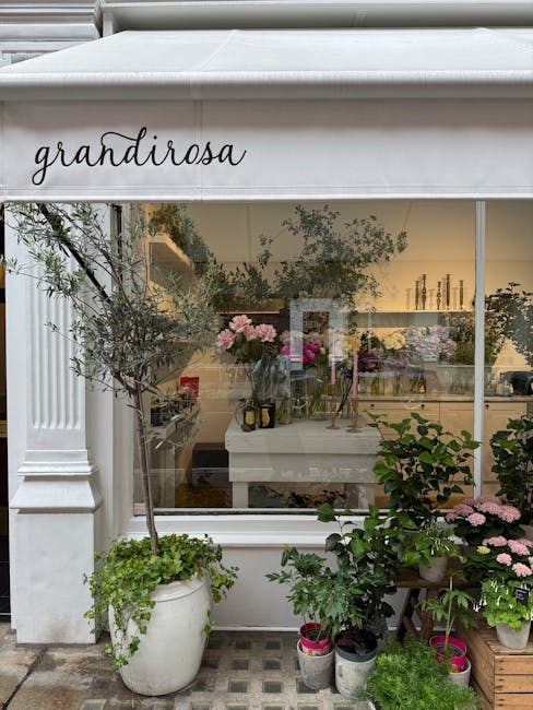 View of a flower shop called 'Grandiroza' through a large glass window, showing an interior with floral arrangements and decorative items. Outside, various potted plants and small trees with lush gree