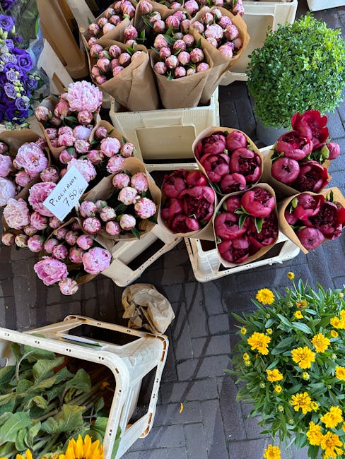 A display of fresh flowers arranged in white crates at an outdoor flower market. There are several bouquets of pink and cream peonies wrapped in brown paper, along with clusters of vibrant pink pomegr