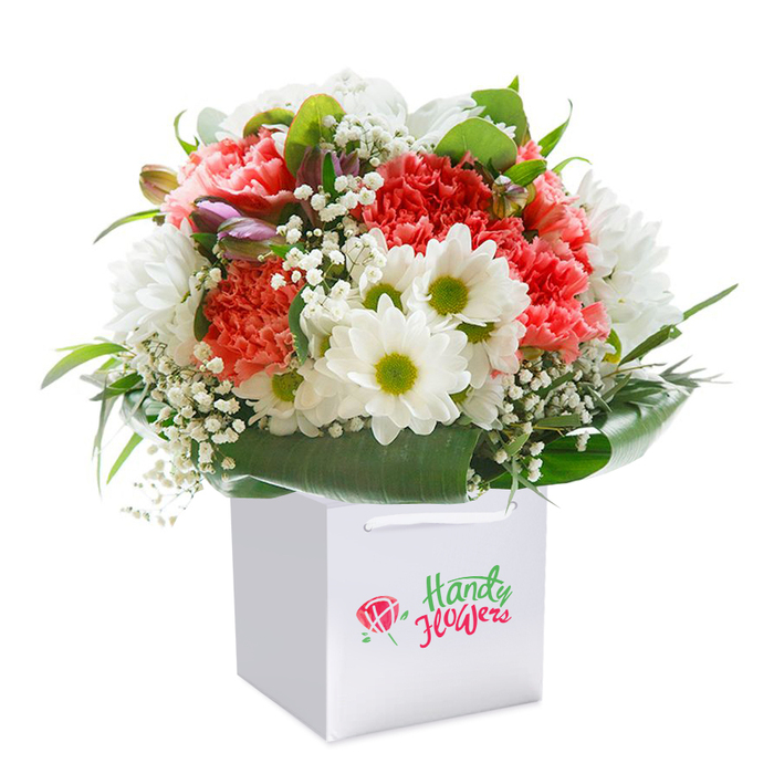 This vibrant bouquet, artfully arranged and elegantly presented in a square white gift bag, echoes the creative spirit found along the streets of Lisson Grove. The centrepiece features a lively mix of coral-pink, peach, and scarlet carnations, their ruffled petals promising lush texture and depth. Among these, daisy-like white chrysanthemums with acid-green centres light up the bouquet with cheer, while ethereal sprays of baby's breath provide a touch of gentle airiness throughout. Subtly positioned purple-tinged buds, likely alstroemeria, introduce an extra layer of complexity and intrigue, drawing the observer's eye to hidden details. Framing the whole composition, abundant broad green foliage forms a natural, glossy collar, reminiscent of the leafy avenues near Lord's Cricket Ground. The sturdy white bag, topped with slim cord handles, enhances the bouquet's ready-to-gift appeal-perfect for quick delivery to a loved one anywhere in the NW8 neighbourhood. Every flower is carefully placed to create a harmonious, full-bodied round shape, demonstrating the expertise and dedication of Florist Lisson Grove. Whether marking a special occasion, offering congratulations, or simply brightening someone's day, this arrangement is a fragrant, fresh ambassador of local floristry talent-ready for same-day delivery throughout Lisson Grove.