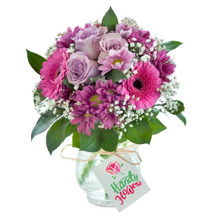 Set against a bright white backdrop, this meticulously curated Lisson Grove floral arrangement features a lush cluster of soft lavender roses at its core, each petal unfurling in a way that suggests quiet elegance and freshness. Flanking the roses, exuberant Gerbera daisies in bold fuchsia pink draw the eye with their vibrant faces and dramatic deep centers, evoking the lively mood of Church Street market. Rich magenta chrysanthemums, shaped like miniature pompons and brushed with hints of plum, provide textural contrast and depth, their radiating petals alluding to the intricate beauty found in local gardens. Sprays of cloud-like baby's breath dot the bouquet, catching the light and offering an airy contrast to the denser, brightly colored blooms. The composition is harmonized by sprigs of deep green foliage, whose glossy surfaces reflect the natural beauty lining Lisson Grove's boulevards. The flowers are expertly arranged in a clear glass vase adorned with a subtly crackled surface, lending a handcrafted feel. A natural raffia bow, tied loosely around the neck of the vase, supports a simple white tag waiting to carry personal words for any occasion. This bouquet, crafted by the florists of Florist Lisson Grove, is a testament to both elegance and joy, ideal for delivery across the local area to brighten birthdays, commemorate milestones, or express heartfelt thanks.