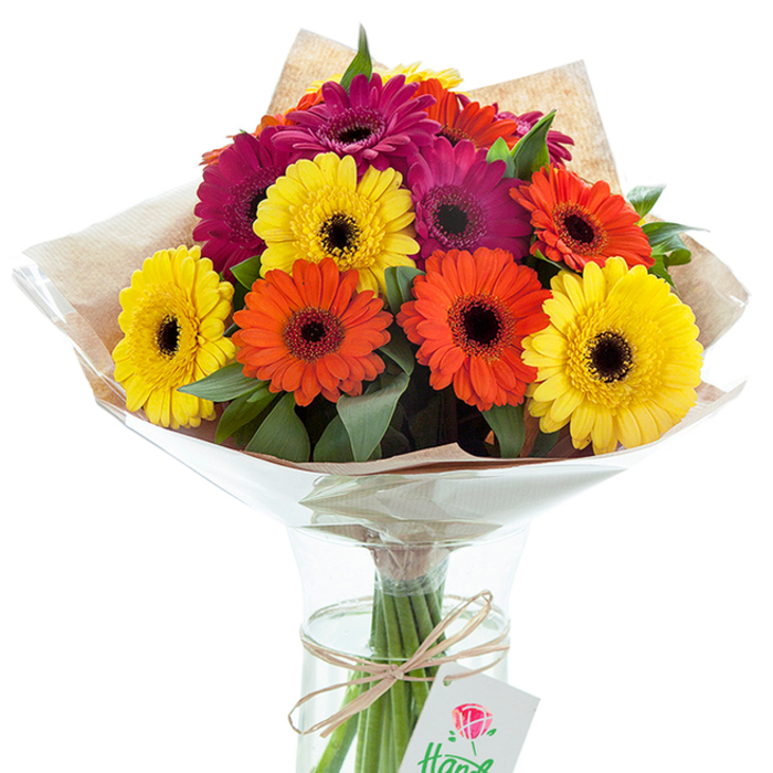 A centerpiece worthy of any Lisson Grove celebration, this generous bouquet of Gerbera daisies is a feast for the senses. Arranged by a local floral artisan, the bouquet features a harmonious balance of striking yellow, bold orange, and deep fuchsia blooms-each daisy a perfect circle radiating from a dark, distinctive center that brings drama and dimension. Lush greenery is interspersed throughout, creating subtle contrast and soft, undulating texture reminiscent of the verdant corners of Paddington Green or the leafy serenity of Regent's Park. The hand-finished wrapping combines tactile, light brown kraft paper-speaking to a rustic, bespoke sensibility-with a layer of gently crinkled clear cellophane, together creating a sophisticated, layered profile that protects and enhances. Resting in a clear glass vase filled with sparkling water, the bouquet's visible stems provide an organic, transparent foundation for the vibrant arrangement above. A piece of natural raffia twine, tied in a simple but decorative bow at the vase's lip, holds a crisp white gift tag featuring a classic red rose illustration-symbolizing the attention to detail that defines Florist Lisson Grove's approach. Whether sent to mark an occasion, brighten an apartment along Lisson Grove, or serve as a gesture of heartfelt thanks, this bouquet speaks of abundance, celebration, and the gentle artistry found within the NW8 community.